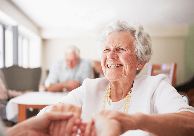 Fröhliche Seniorin mit weißem Haar und weißer Kleidung sitzt in einem hellen Raum und hält lächelnd die Hände einer anderen Person. Im Hintergrund sind weitere ältere Menschen in einer freundlichen Gemeinschaftsatmosphäre zu sehen.